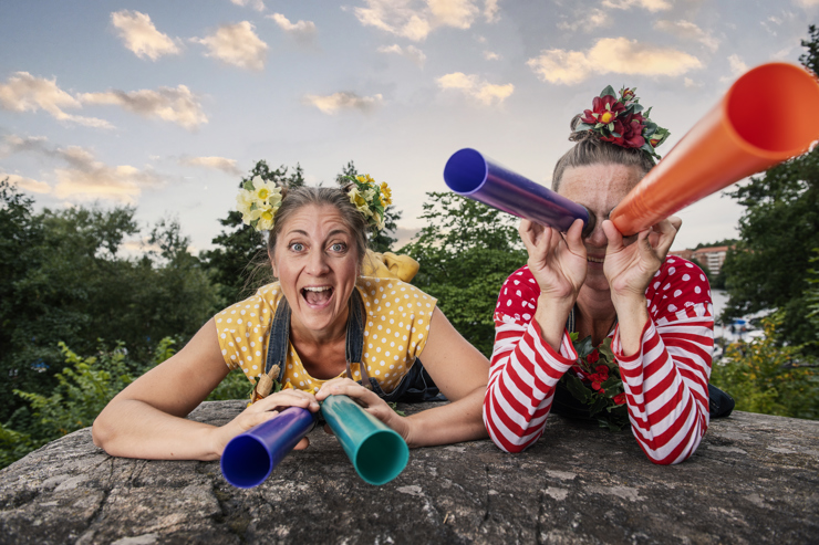 Två färgglada kvinnor ligger och spanar - den ena med kikare för ögonen, den andra med kikare på marken.