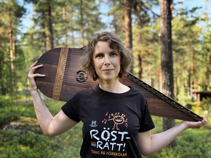 Foto av Mirja Palo som står i skogen och håller sitt instrument - en kantele.
